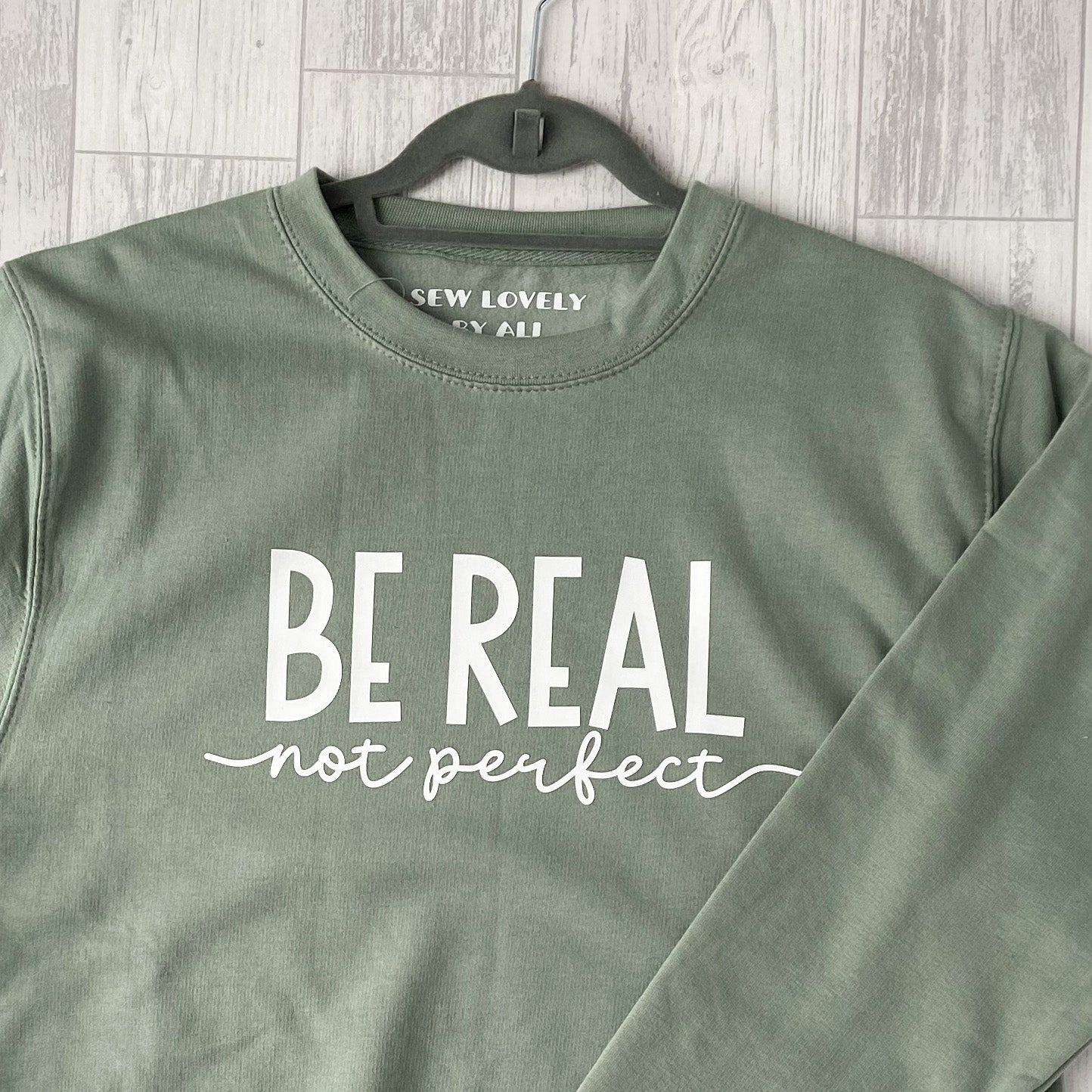 Dusty green crew neck sweatshirt with white wording that reads 'Be Real Not Perfect'.