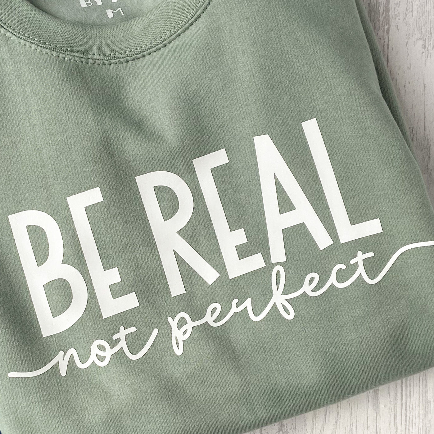 Dusty green crew neck sweatshirt with white wording that reads 'Be Real Not Perfect'.